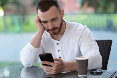 Açık kafede akıllı telefon kullanan, kağıt bardaklı yakışıklı bir adam.