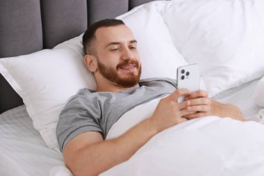 Yatakta cep telefonu kullanan gülümseyen adam.