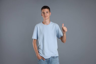 Açık mavi tişört giyen ve gri arka planda baş parmak gösteren genç çocuk.