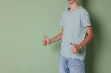 Açık gri tişört giyen ve yeşil arka planda baş parmak gösteren genç çocuk, yakın plan. Metin için boşluk