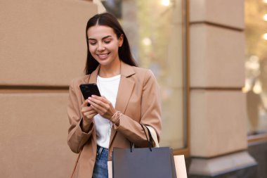 Açık havada akıllı telefon kullanan alışveriş çantalı mutlu bir kadın.