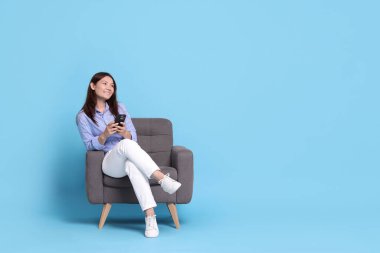 Akıllı telefonlu gülümseyen kadın açık mavi arka planda koltukta dinleniyor. Metin için boşluk