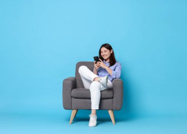 Akıllı telefonlu gülümseyen kadın açık mavi arka planda koltukta dinleniyor.