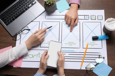 UI tasarımı geliştiriliyor. Tasarımcılar, ahşap masadaki kağıt üzerinde web sitesi çerçevesi oluşturuyorlar, üst görünüm