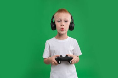 Kulaklıklı şirin bir çocuk yeşil arka planda kontrolörle video oyunu oynuyor.