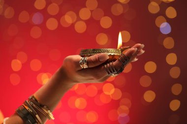 Diwali kutlaması. Kırmızı arka planda yanan diya lambasını tutan bulanık ışıklı kadın, yakın plan.