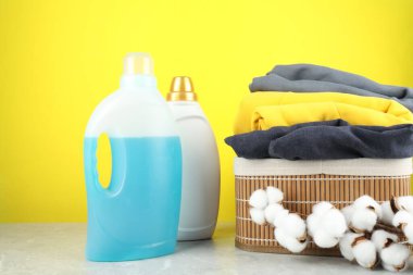 Şişelerde çamaşır deterjanı, pamuk çiçekleri ve gri mermer masada sarı arka planda giysiler.