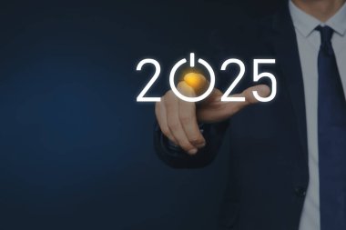 Yeni yıl hedefleri. İş adamı, koyu mavi arkaplana karşı sanal ekranda sıfır yerine güç düğmesi ile 2025 tuşuna basıyor.