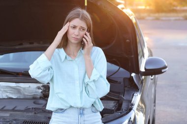 Stresli bir kadın dışarıda kırık bir arabanın yanında telefonla konuşuyor.