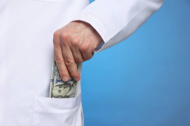 Yozlaşma konsepti. Doktor cebine dolar banknotları koyuyor mavi arka planda, yakın plan. Metin için boşluk