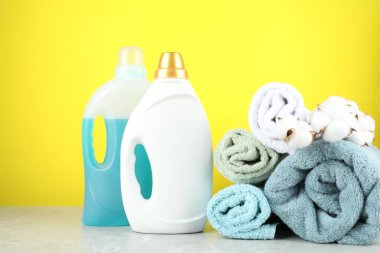 Çamaşır deterjanı şişelerde, pamuk çiçekleri ve havlular gri mermer masada sarı arka planda.