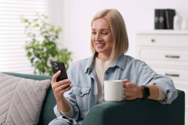 Gülümseyen orta yaşlı kadın bir fincan sıcak içecekle evde akıllı telefon kullanıyor.