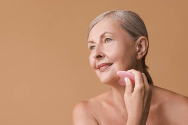 Güzel bir kadın bej arka planda gua sha aracı ile yüz masajı yapıyor, mesaj için boşluk