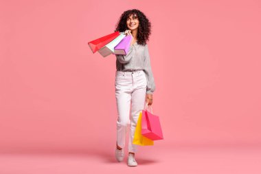 Pembe arka planda renkli alışveriş torbalarıyla gülümseyen kadın.