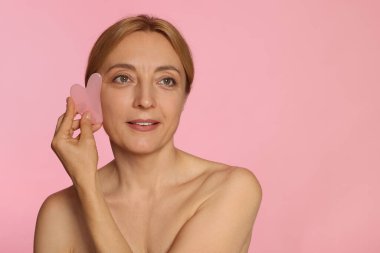 Gülümseyen kadın pembe arka planda gua sha aletiyle yüz masajı yapıyor. Metin için boşluk
