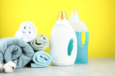 Çamaşır deterjanı şişelerde, pamuk çiçeği ve havlular gri mermer masada sarı arka planda.