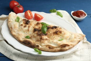 Fesleğenli, domatesli ve mavi ahşap masadaki soslu leziz calzone, yakın plan.