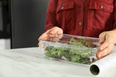 Kadın, mutfaktaki beyaz masada salata ile cam kabın üzerine plastik yiyecek örtüsü koyuyor.