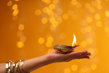 Diwali kutlaması. Elinde yanan diya lambası olan turuncu arka planda bulanık ışıkları olan bir kadın, yakın plan.