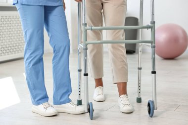 Klinikte yürüyen çerçeveli yaşlı bir kadının yanında hemşire, yakın plan.