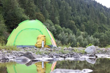 Kamp çadırı, sırt çantası ve termosu dağlarda nehir kenarında taşların üzerinde.