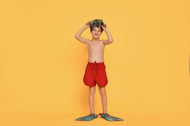 Sahilde dalış maskesi ve turuncu arka planda paletleri olan küçük bir çocuk.