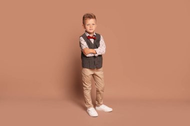 Kahverengi arka planda şık giyinen küçük bir çocuğun tam boy portresi.