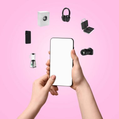 İnternet alışverişi. Elinde boş ekranlı cep telefonu tutan kadın pembe arka planda farklı ev aletleri ve cihazlarla çevrili.