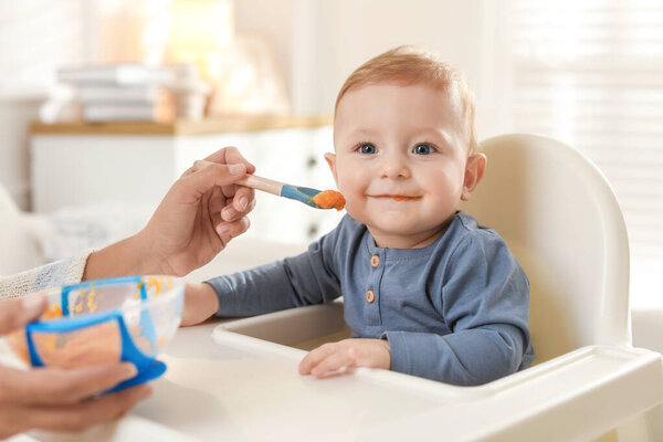 Mother feeding her cute little baby in high chair at home, closeup