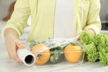 Beyaz mermer masada plastik gıda ambalajını taze sebzelerle kaplayan kadın, yakın plan.