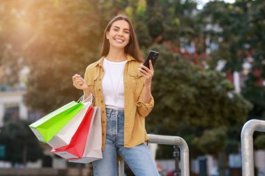 Açık havada akıllı telefon kullanan renkli alışveriş çantalı mutlu bir kadın.