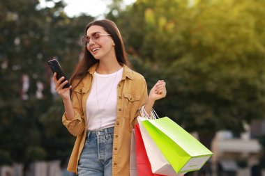 Açık havada akıllı telefon kullanan renkli alışveriş çantalı mutlu bir kadın.
