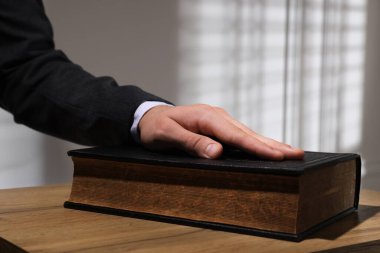 Man taking oath with his hand on Bible at wooden table, closeup clipart