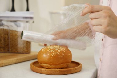 Woman putting plastic food wrap over fresh bun at white table in kitchen, closeup clipart