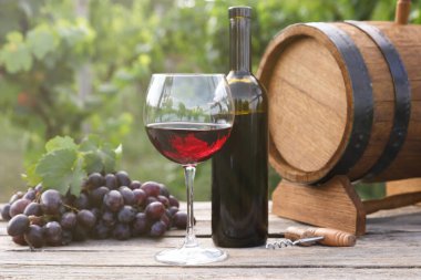 Kırmızı şarap şişede, cam, fıçı ve üzüm dışarıda, ahşap masada.