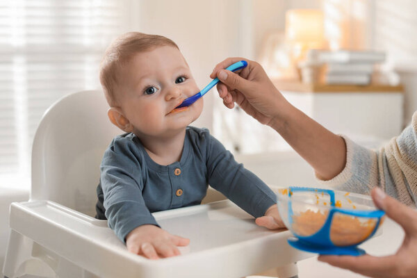 Mother feeding her cute little baby in high chair at home, closeup