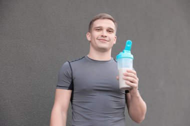 Gri duvarın yanında protein içeceği sallayan atletik bir adam.