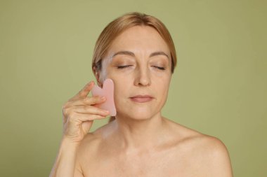 Yeşil arka planda gua sha aracıyla yüz masajı yapan güzel bir kadın.
