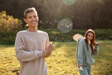 Güneşli bir günde parkta badminton raketleri ve mekik aleti olan genç bir adam ve bir kadın, seçici bir odak noktası.