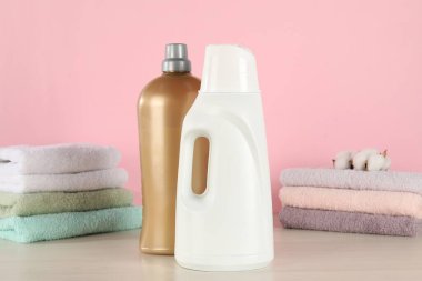 Çamaşır deterjanı, kep, temiz havlular ve ahşap masada pembe arka planda duran pamuk çiçekli şişeler.