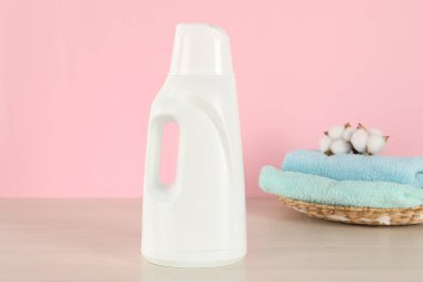 Çamaşır deterjanı şişesi, temiz havlular ve ahşap masanın üzerinde pembe arka planda duran pamuk çiçekleri.
