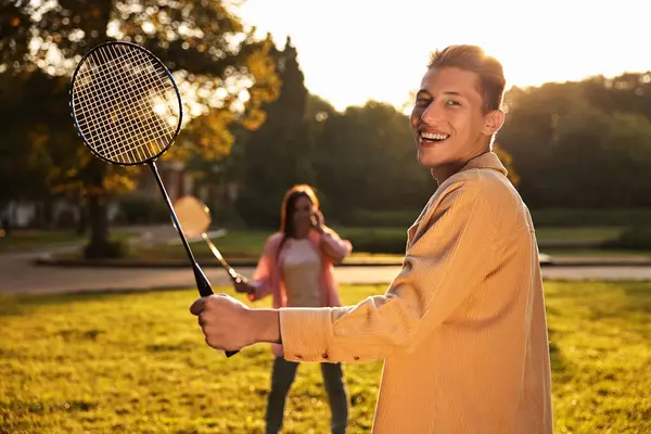 Genç adam ve kadın güneşli bir günde parkta badminton oynuyorlar.