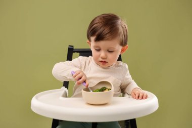 Küçük şirin bebek, zeytin arka planında yüksek sandalyedeki kaseden sağlıklı yemek yiyor.