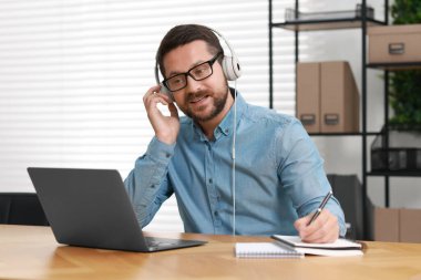 Kulaklıklarda tercüman. İçerideki ahşap masada bilgisayarla görüntülü konuşma yaparken not alıyor.