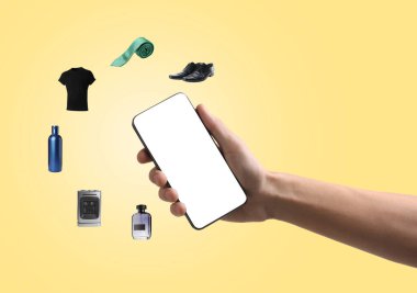 Sarı arka planda online alışveriş için cep telefonu kullanan bir adam, yakın plan. Farklı mallar etrafta uçuşuyor. Boş ekranlı aygıt