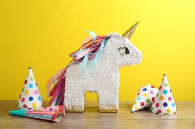 Tek boynuzlu at şeklindeki güzel pinyatalar, parti konileri ve ahşap masada sarı arka planda duran patlatıcılar.