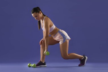 Kadın, mor arka planda dambıllarla egzersiz yapıyor.