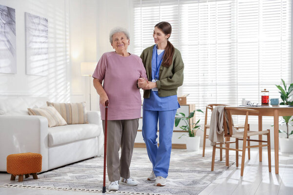 Caregiver assisting senior woman with walking cane indoors. Home health care service