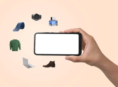 Bej arka planda online alışveriş için cep telefonu kullanan bir adam, yakın plan. Farklı mallar etrafta uçuşuyor. Boş ekranlı aygıt