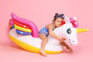 Cute little girl in swimsuit with unicorn pool float on light pink background clipart